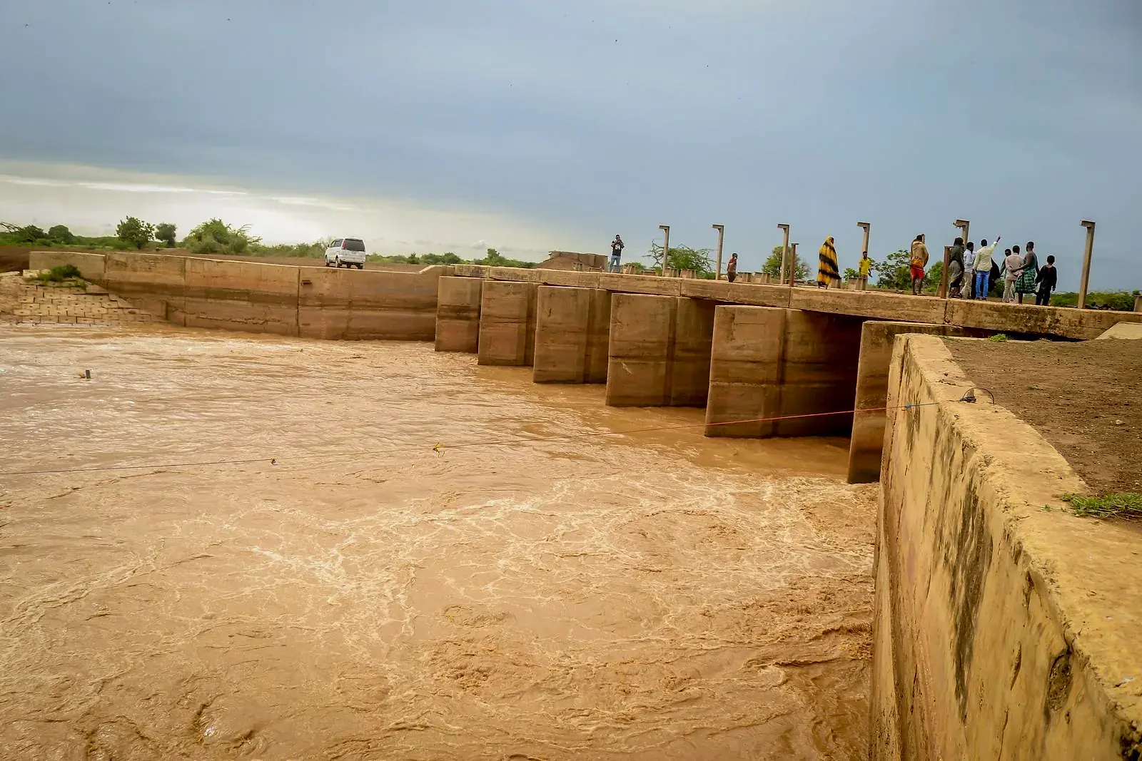 A concrete river barrage with brown floodwater surging through every gate, and a line of people watching from the crossing above.