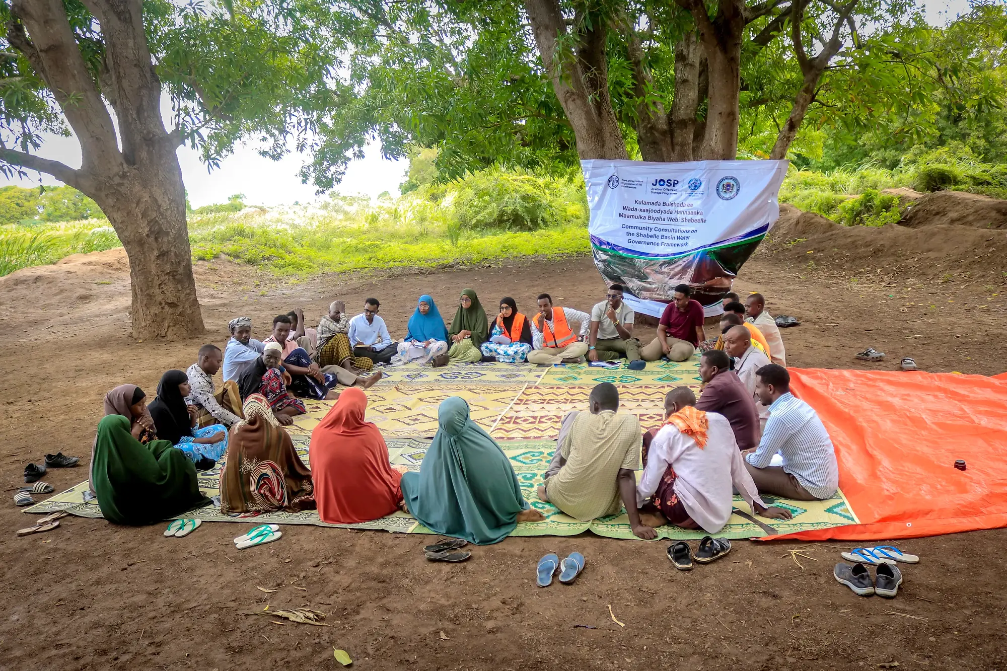 A large community consultation seated on mats beneath a project banner strung between two trees.