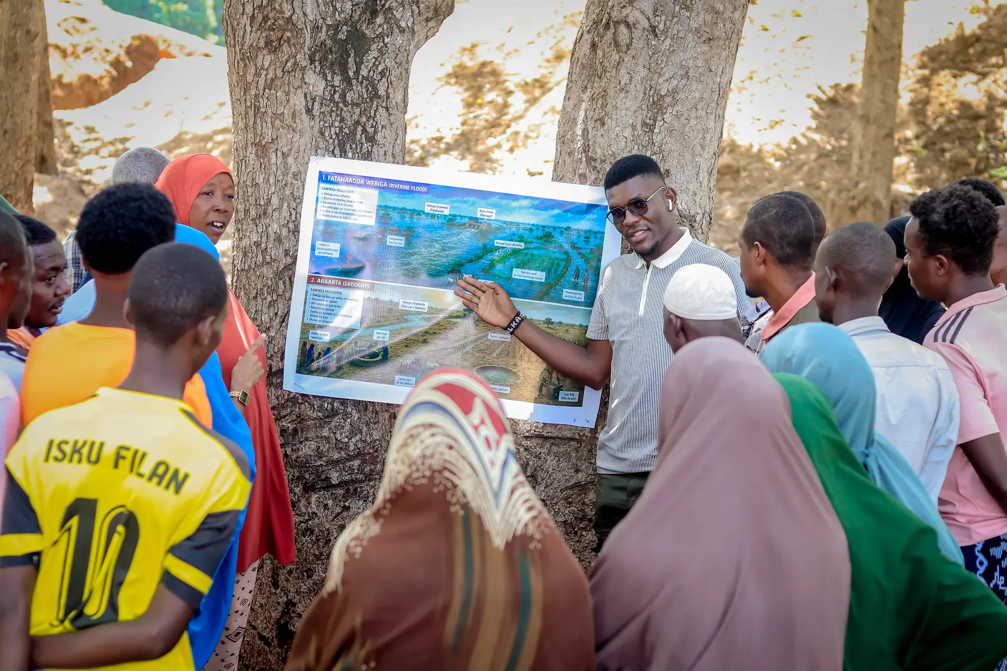 A facilitator holds a large printed map board and points to a feature on it while participants watch.