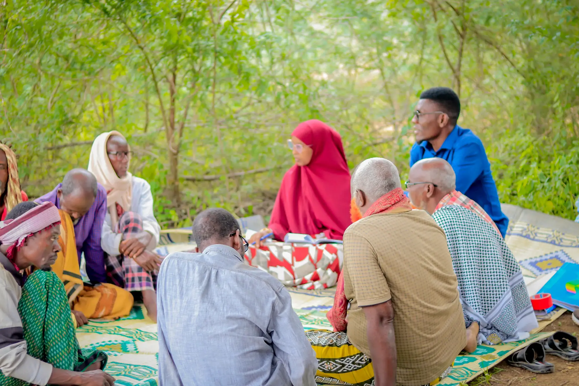 Women and older men seated together on mats in the shade, listening.