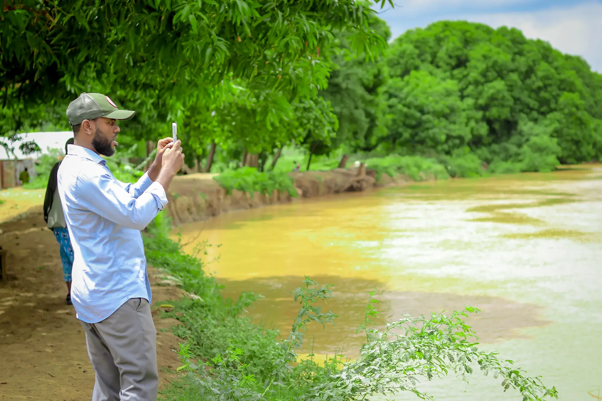 A man stands on the bank photographing a silted, discoloured channel on his phone.