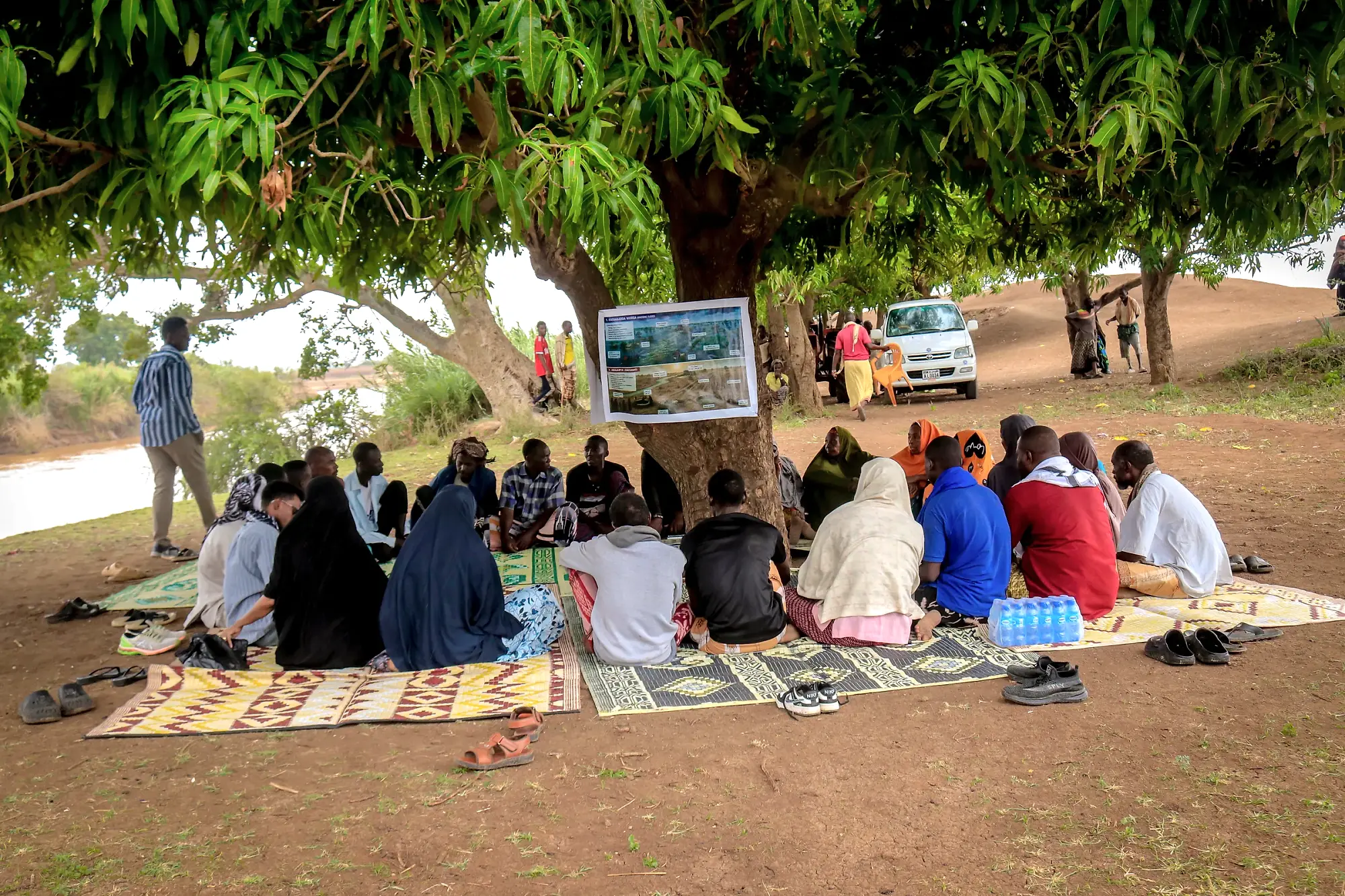 A community session seated on mats in a wide circle under a mango tree, a map board propped against the trunk.